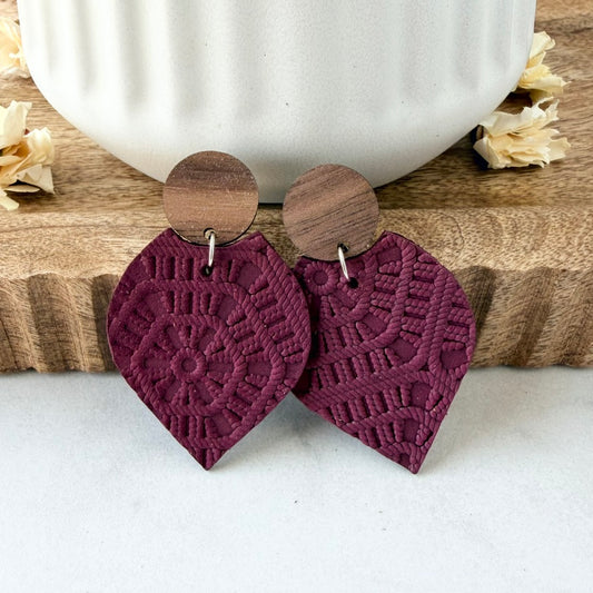A pair of bohemian-style dangle earrings with textured, embossed burgundy petal-shaped leather bottoms and circular wooden studs, displayed on a wooden board with a white ceramic mug in the background.