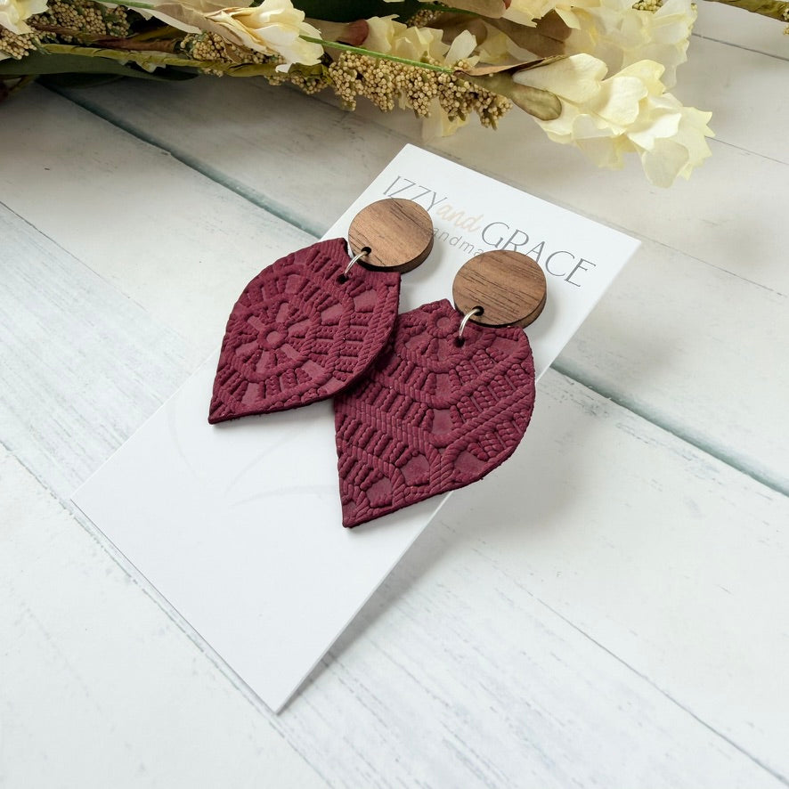 A pair of dangling earrings displayed on a white card. Each earring has a round natural wood top and a larger petal-shaped drop made of burgundy embossed leather with a lace-like pattern.