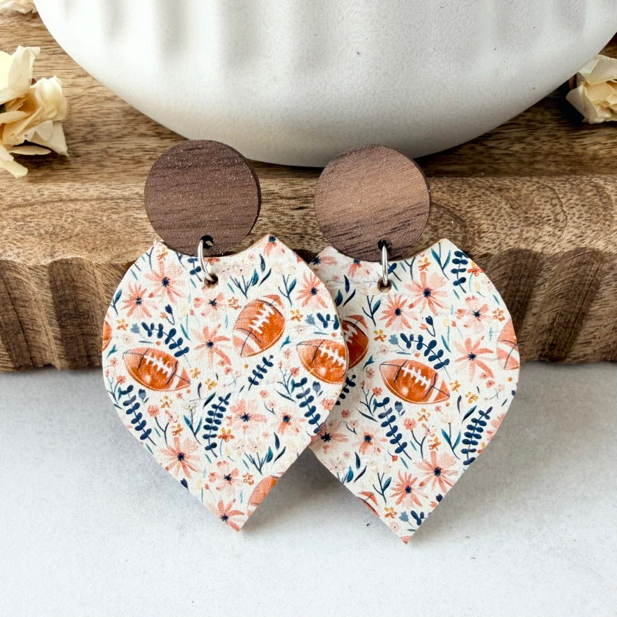 A pair of petal-shaped drop earrings with a floral and football print, hanging from round wooden studs on a rustic wooden surface with a white ceramic bowl in the background.