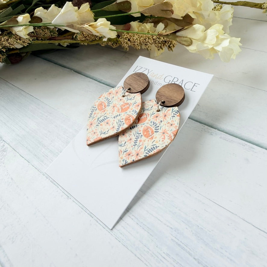 A pair of drop earrings displayed on a white card. Each earring has a round dark wood top and a petal-shaped lower section covered in a pink and, blue floral pattern with brown footballs.