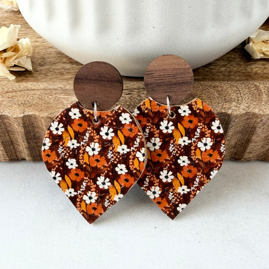 A pair of petal-shaped drop earrings with a brown wood circular top and a patterned lower section featuring orange, white, and dark brown floral motifs, displayed on a wooden block with a white ceramic bowl in the background.