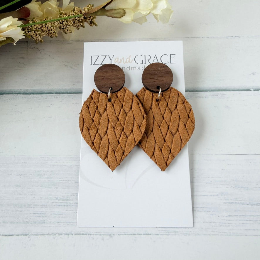 A pair of handmade dangle earrings displayed on a white card. The earrings feature textured, woven mustard leather petal-shaped drops with round dark brown wooden studs.