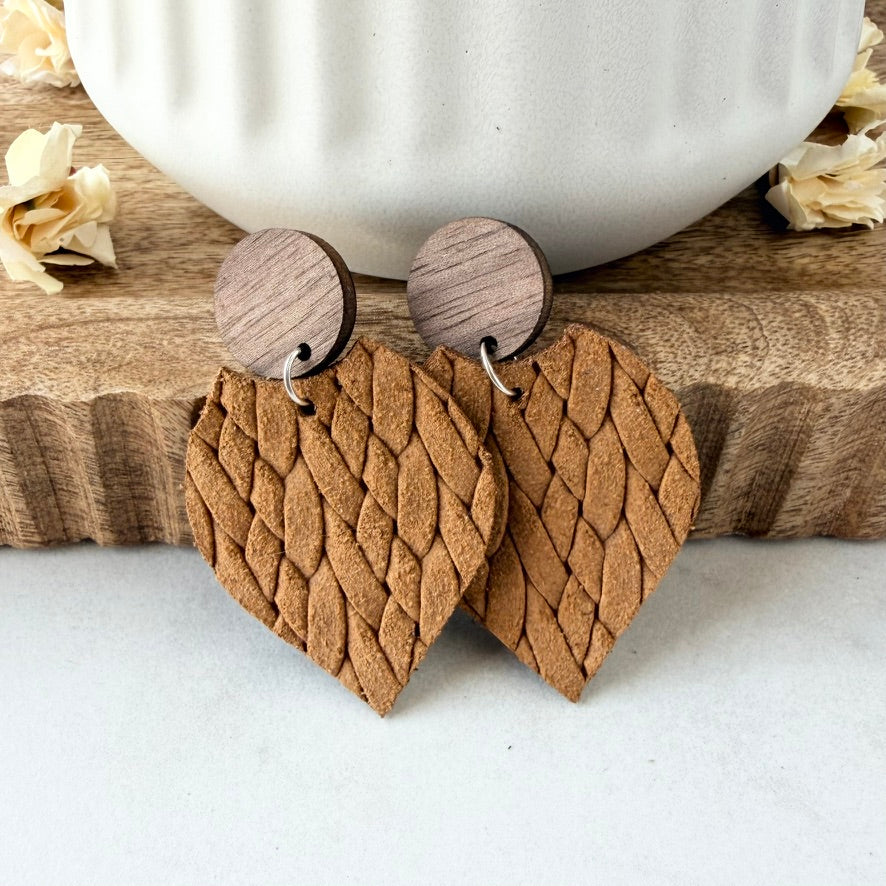 petal-shaped wooden drop earrings with a textured, braided leather pattern in a warm mustard, featuring circular wood studs, displayed on a wooden block with a white ceramic mug in the background.