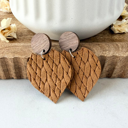 petal-shaped wooden drop earrings with a textured, braided leather pattern in a warm mustard, featuring circular wood studs, displayed on a wooden block with a white ceramic mug in the background.