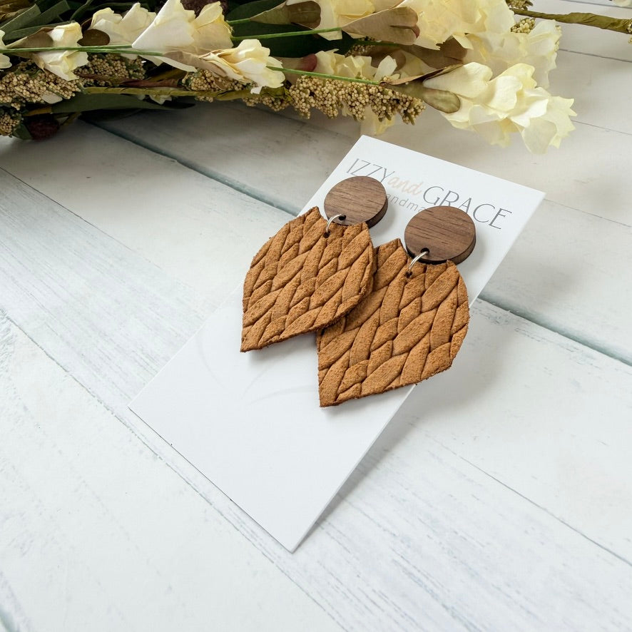 A pair of textured mustard petal-shaped drop earrings with round wooden studs at the top, displayed on a white card labeled 'Izzy and Grace' on a white woo surface with dried flowers in the background.