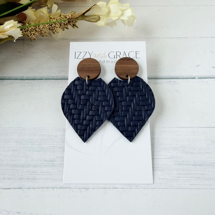 A pair of dark navy woven leather drop earrings with round wooden studs, displayed on a white card labeled 'IZZYand GRACE' on a white wood background with dried flowers in the corner.