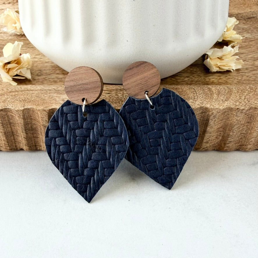 A pair of handmade drop earrings featuring dark navy blue woven leather petal bottom and round natural wood studs, displayed on a wooden block with a white ceramic mug in the background.
