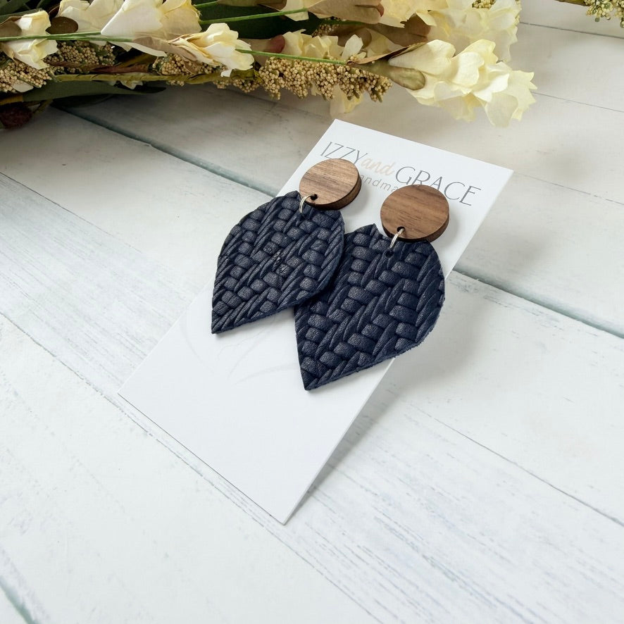 A pair of textured dark navy blue woven drop earrings with round wooden studs, displayed on a white branded card that reads 'Izzy and Grace'. The card is placed on a white wooden surface with dried flowers in the background.