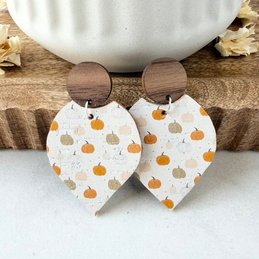 A pair of dangle earrings with round dark wood tops and petal-shaped lower pieces printed with small orange, beige, and gray pumpkins on a white background, styled on a wooden surface with a white ceramic bowl.