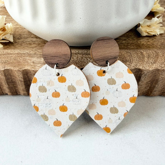 A pair of dangle earrings with round dark wood tops and petal-shaped lower pieces printed with small orange, beige, and gray pumpkins on a white background, styled on a wooden surface with a white ceramic bowl.