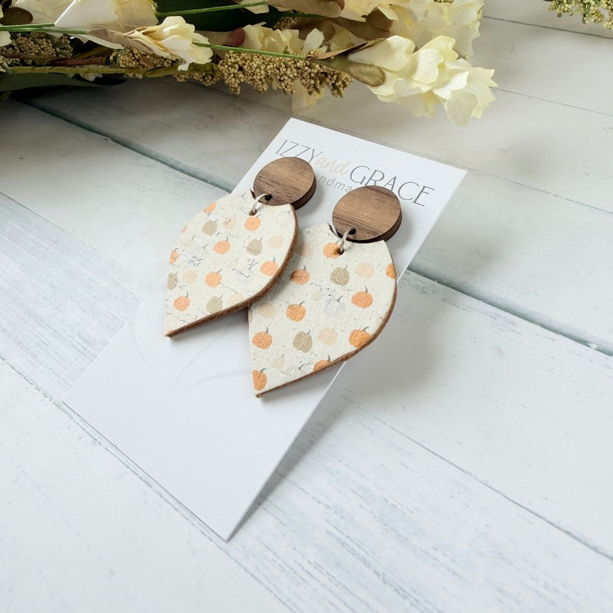 A pair of wooden dangle earrings with a cream-colored fall-themed pattern of orange and beige pumpkins, hanging from round brown wooden studs. The earrings are displayed on a white card labeled 'LIZZY IN GRACE' on a light gray wooden surface with dried flowers in the background.