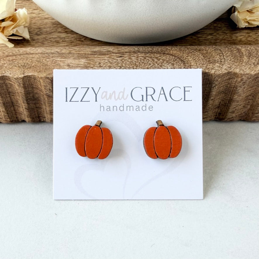 A pair of orange pumpkin-shaped stud earrings displayed on a white card labeled 'IZZY and GRACE handmade,' placed on a wooden surface with dried flowers and a white bowl in the background.