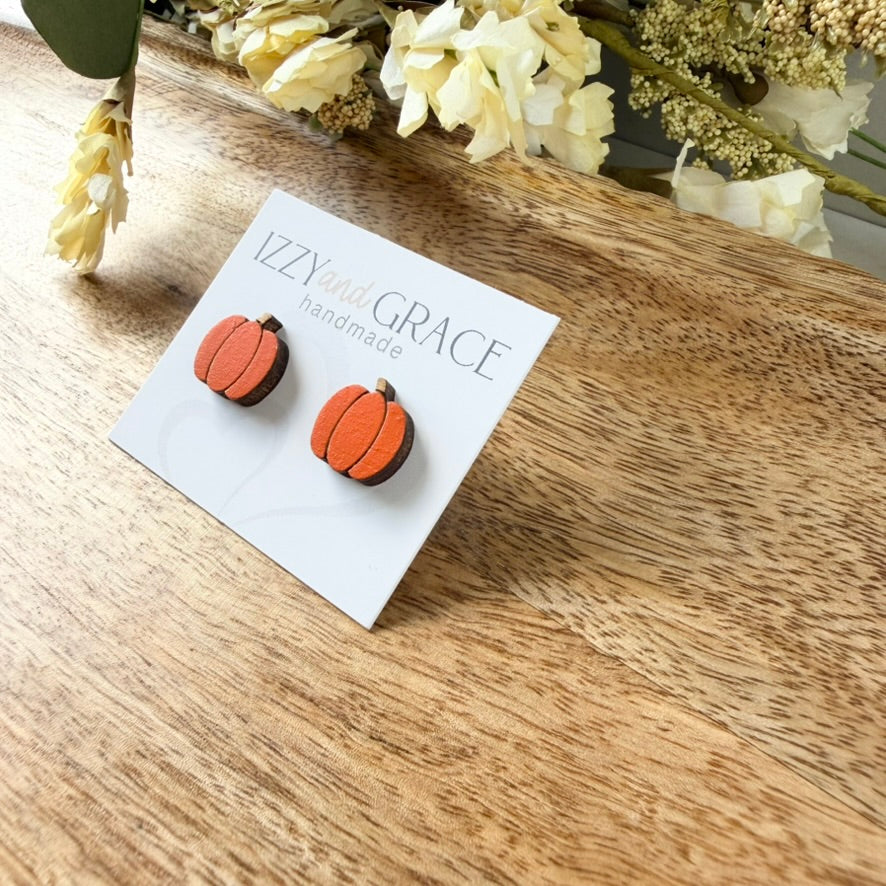 A pair of orange and brown pumpkin-shaped stud earrings displayed on a white card labeled 'Izzy and Grace handmade,' placed on a wooden surface with dried flowers in the background.
