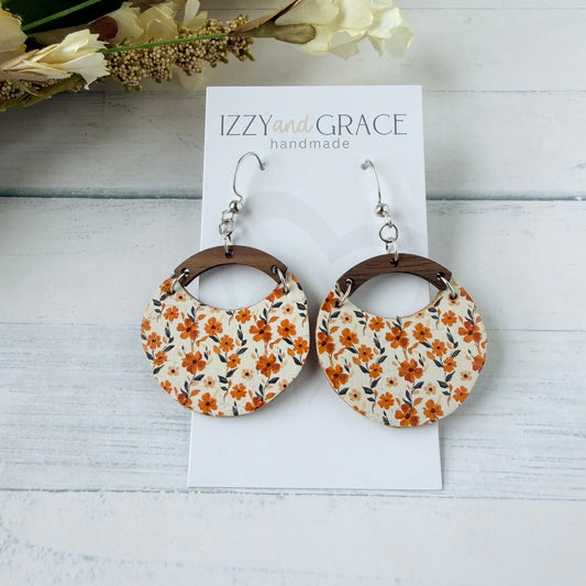 A pair of handmade dangle earrings displayed on a white card labeled 'IZZY and GRACE handmade'. The earrings feature a circular design with a cream background, orange and yellow floral pattern, and brown wooden arches, hanging from silver fishhook wires.