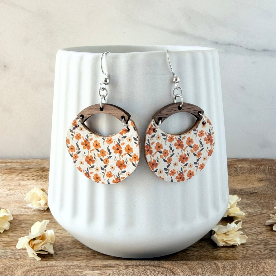 A pair of circular drop earrings with a white background and orange floral pattern, featuring dark brown wooden hoops and silver fishhook wires, displayed on a white ribbed ceramic vase.