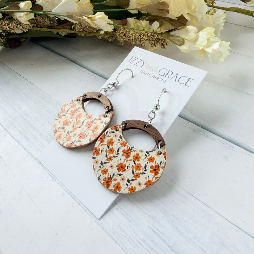 A pair of handmade dangle earrings displayed on a white card labeled 'Izzy and Grace Handmade.' The earrings have circular floral panels with orange and yellow flowers on a cream background, brown wooden arches, and silver hooks.