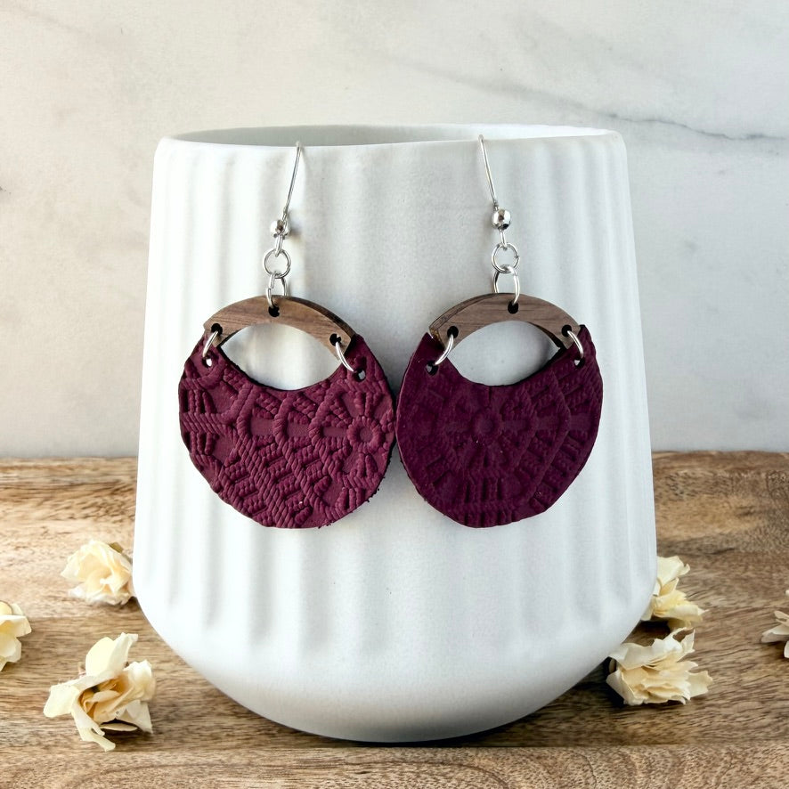 A pair of handmade dangle earrings with textured deep burgundy circular discs and natural wood arches, hanging in front of a white ceramic vase on a wooden surface.