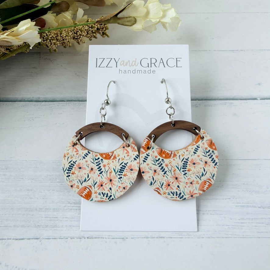 A pair of handmade dangle earrings displayed on a white card labeled 'IZZY and GRACE handmade'. The earrings feature circular leather with a floral pattern in orange, blue, white, and footballs connected by brown wooden arches and hanging from silver fishhook wires.