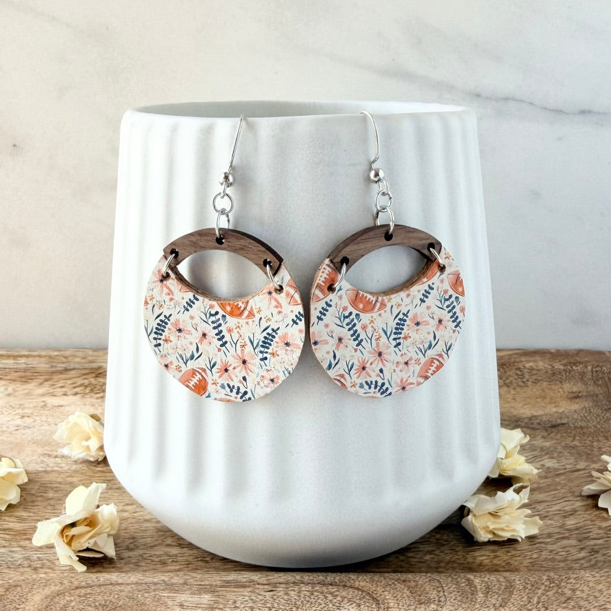 A pair of dangling hoop earrings with a floral print, displayed on a white ribbed ceramic vase. The earrings feature a pattern of orange, pink, and blue flowers with brown footballs on a white background, with dark brown wooden arches and silver hooks.