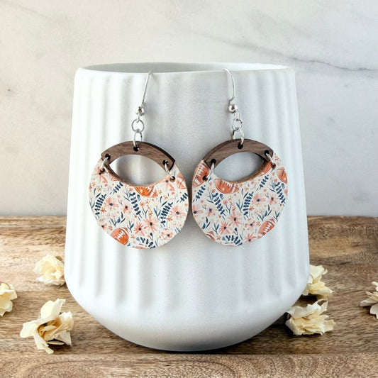 A pair of dangling hoop earrings with a floral print, displayed on a white ribbed ceramic vase. The earrings feature a pattern of orange, pink, and blue flowers with brown footballs on a white background, with dark brown wooden arches and silver hooks.