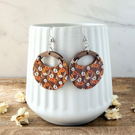 A pair of large circular drop earrings with a brown, orange, and white floral pattern, hanging from silver fishhook wires. They are displayed on a white ribbed ceramic vase set on a wooden surface with dried flowers.
