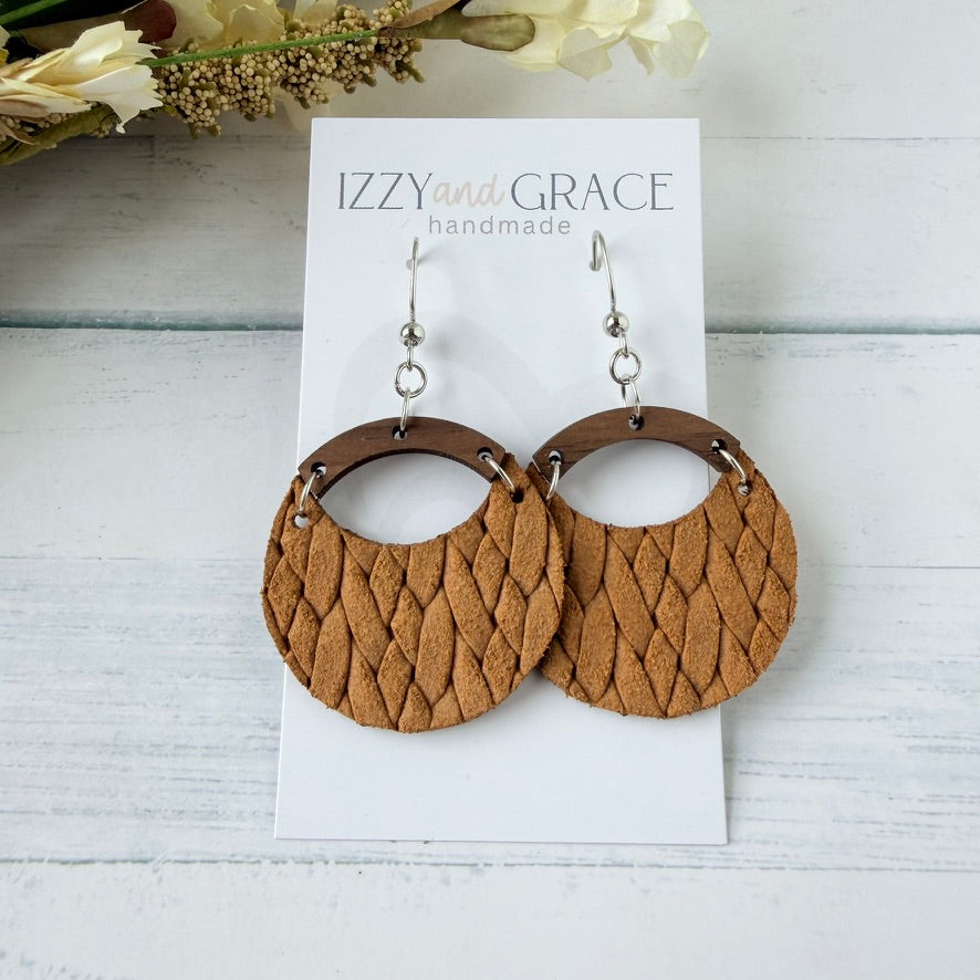 A pair of large circular dangle earrings with textured mustard woven leather and dark brown wooden arches, displayed on a white card labeled 'IZZY and GRACE handmade' on a white wood background.