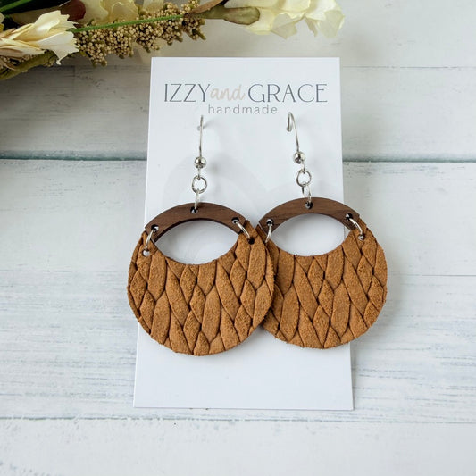 A pair of large circular dangle earrings with textured mustard woven leather and dark brown wooden arches, displayed on a white card labeled 'IZZY and GRACE handmade' on a white wood background.