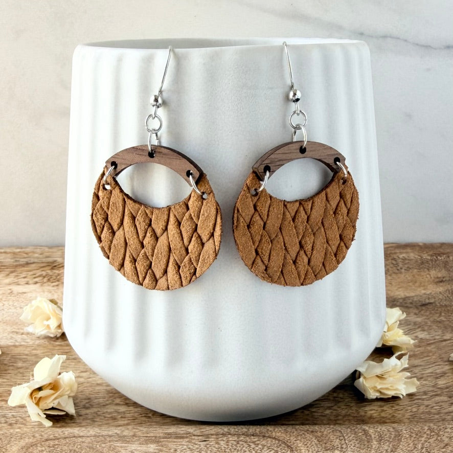 A pair of round dangle earrings with woven mustard leather and dark wood accents, displayed on a white ribbed ceramic vase on a wooden surface.