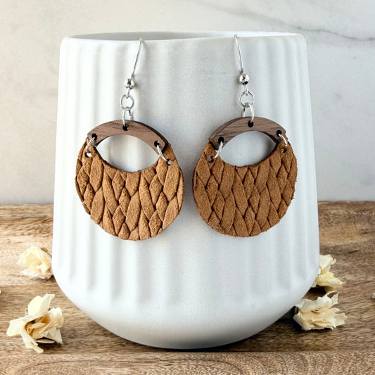 A pair of round dangle earrings with woven mustard leather and dark wood accents, displayed on a white ribbed ceramic vase on a wooden surface.
