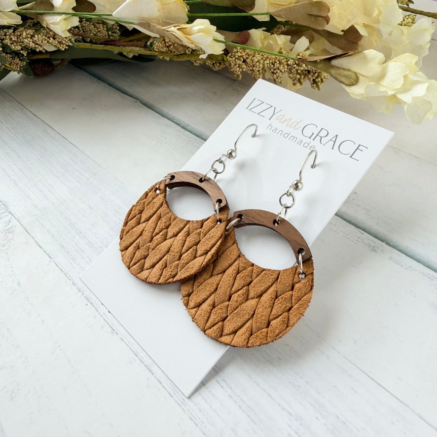 A pair of large, circular bohemian-style dangle earrings made of textured mustard leather with a wooden arch and silver hooks, displayed on a white card labeled 'IZZY and GRACE handmade' on a white wooden surface with dried flowers in the background.