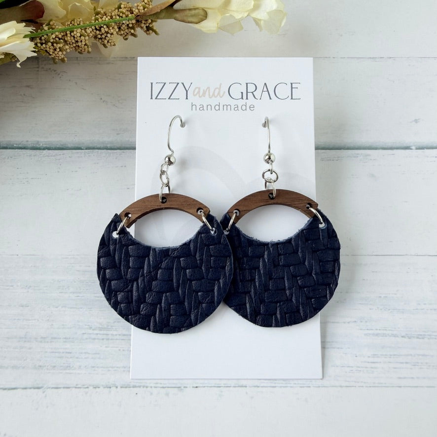 A pair of handmade dangle earrings displayed on a white card labeled 'IZZY and GRACE handmade.' The earrings have dark navy woven circular drops with brown wooden arches and silver fishhook wires, photographed on a light wood surface with dried flowers in the corner.