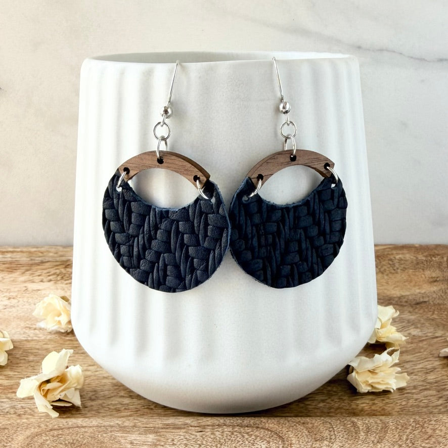 A pair of large oval drop earrings with dark navy woven-texture discs, light brown wooden arches, and silver fishhook wires, displayed on a white ribbed ceramic vase.