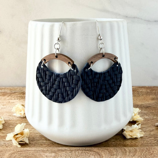 A pair of large oval drop earrings with dark navy woven-texture discs, light brown wooden arches, and silver fishhook wires, displayed on a white ribbed ceramic vase.