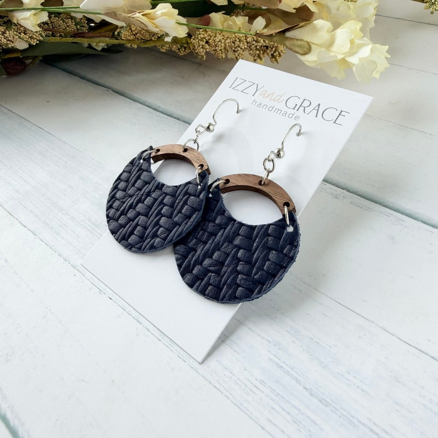 A pair of handmade dangle earrings displayed on a white card. The earrings feature dark navy blue woven leather circular drops with a wood arch connector and silver fishhook wires.