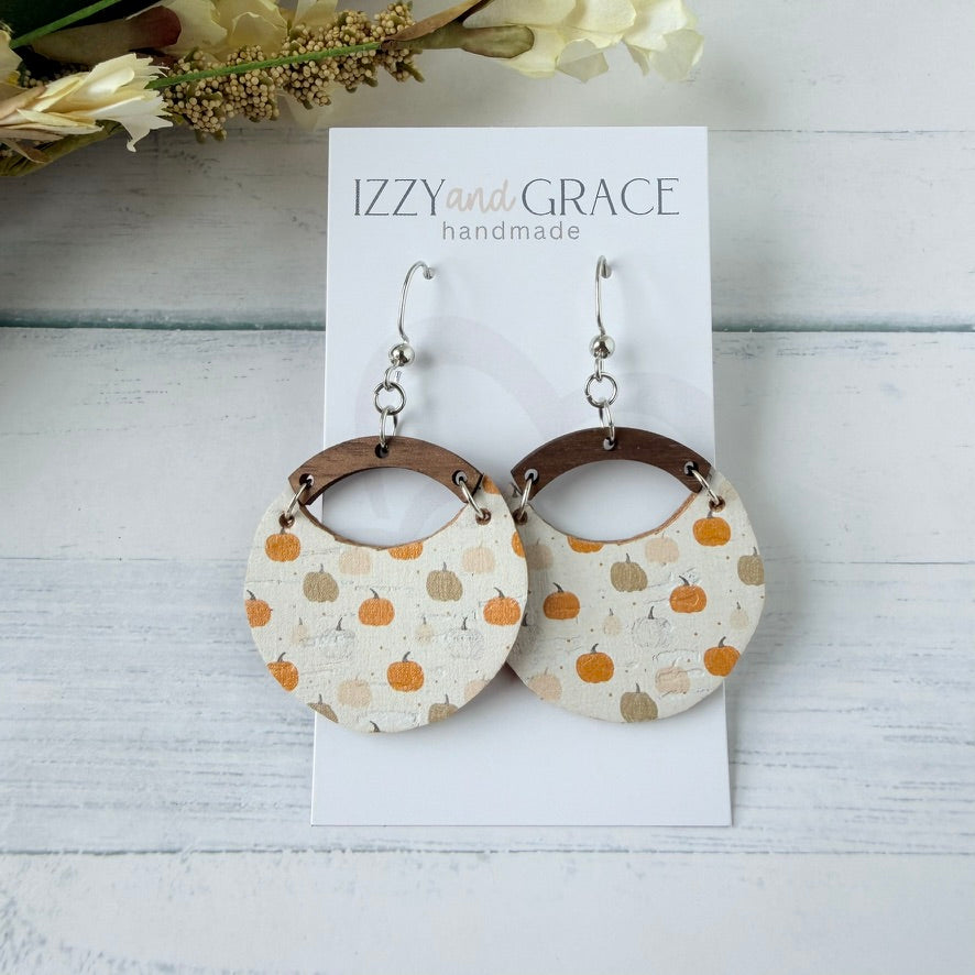 A pair of handmade dangle earrings displayed on a white card labeled 'IZZY and GRACE handmade.' The earrings have silver fishhook wires and circular beige leather bottom with an orange and tan pumpkin pattern, connected by brown wooden arches.