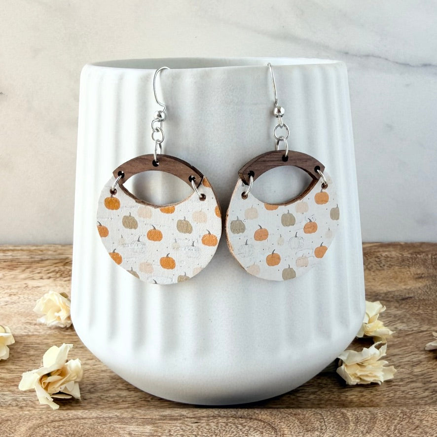 A pair of dangling earrings displayed on a white ribbed ceramic cup. The earrings feature silver fishhook wires, dark brown wooden arches, and round leather bottoms printed with small orange and beige pumpkins.