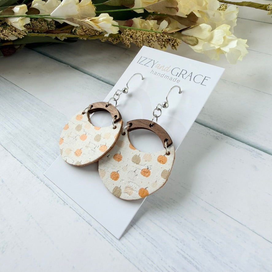 Pumpkin-themed dangle earrings on a white display card. The earrings feature a circular beige and white patterned disc with small orange and brown pumpkins, connected by a brown wooden arch to a silver fishhook ear wire.