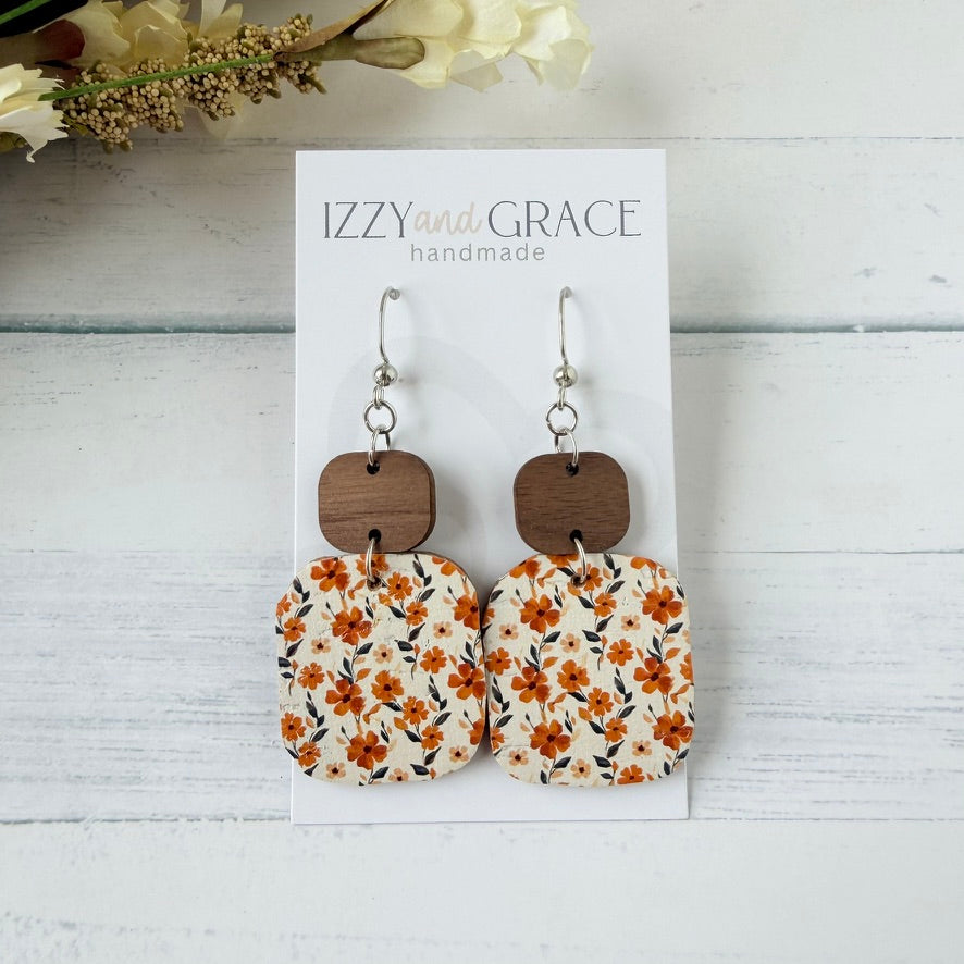 A pair of dangle earrings displayed on a white card labeled 'IZZY and GRACE handmade,' set against a whitewashed wood background with dried flowers in the corner. The earrings have silver fishhook wires, dark brown wooden square connectors, and cream fabric squares with an orange floral print.