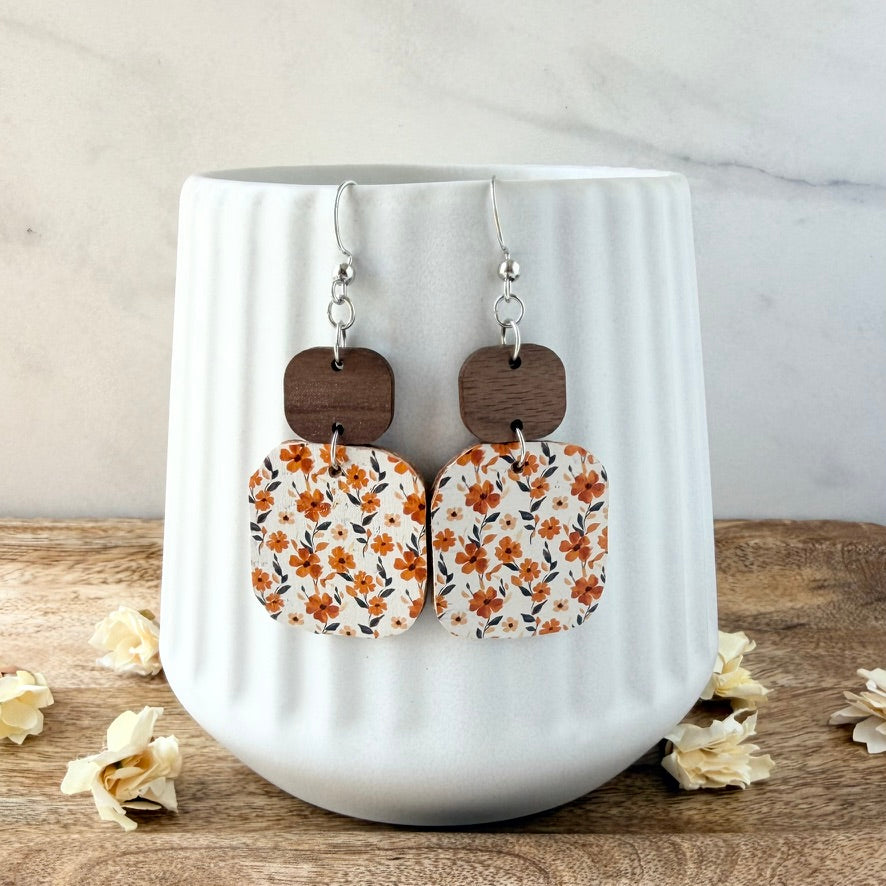 A pair of drop earrings displayed on a white ribbed ceramic vase. The earrings feature silver fishhook wires, dark brown wooden square connectors, and rectangular wooden charms printed with a white background and a warm orange floral pattern.