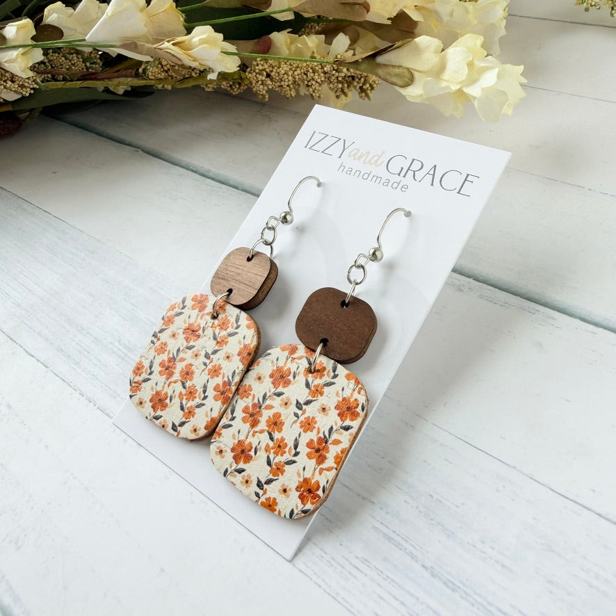 A pair of handmade rectangular drop earrings with a cream background and orange floral print, attached to silver fishhook wires and a small brown wooden top, displayed on a white card labeled 'IZZY and GRACE handmade' on a white wood surface.