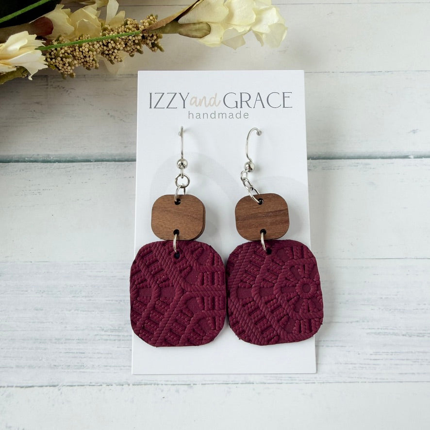 A pair of handmade drop earrings displayed on a white card labeled 'IZZY and GRACE handmade.' Each earring has a silver fishhook wire, a small rounded square wooden charm, and a larger textured rectangular drop in deep burgundy.