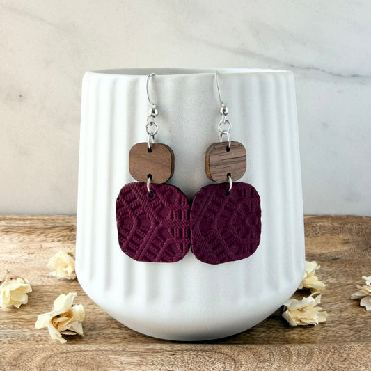 A pair of handmade dangle earrings with silver fishhook wires, square brown wood charms, and textured deep burgundy square drops, displayed on a white ribbed ceramic cup with dried flowers on a wooden surface.