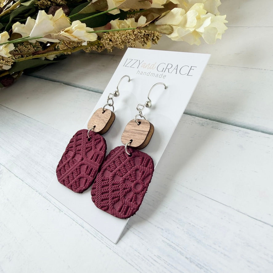 A pair of handmade dangle earrings displayed on a white card. The earrings feature textured burgundy leather rectangular drops with a wooden connector and silver-tone hooks.