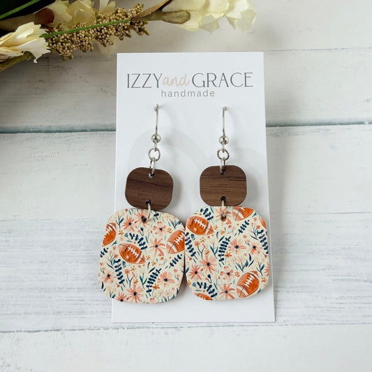 A pair of dangling earrings displayed on a white card labeled 'IZZY and GRACE handmade.' The earrings have silver hooks, dark brown wooden square tops, and floral patterned drop charms with football motifs on a light background.