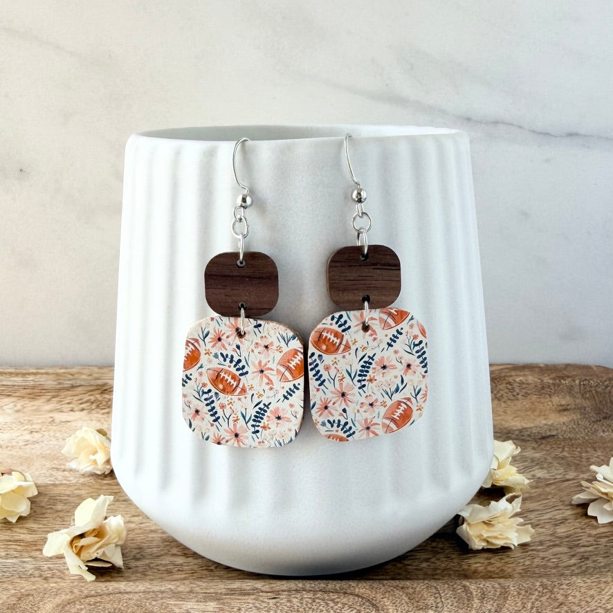 A pair of dangling earrings displayed on a white ribbed ceramic mug. The earrings feature silver fishhook wires, dark brown wooden square charms, and rectangular fabric charms with a floral and football pattern in pink and blue.