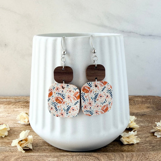 A pair of dangling earrings displayed on a white ribbed ceramic mug. The earrings feature silver fishhook wires, dark brown wooden square charms, and rectangular fabric charms with a floral and football pattern in pink and blue.