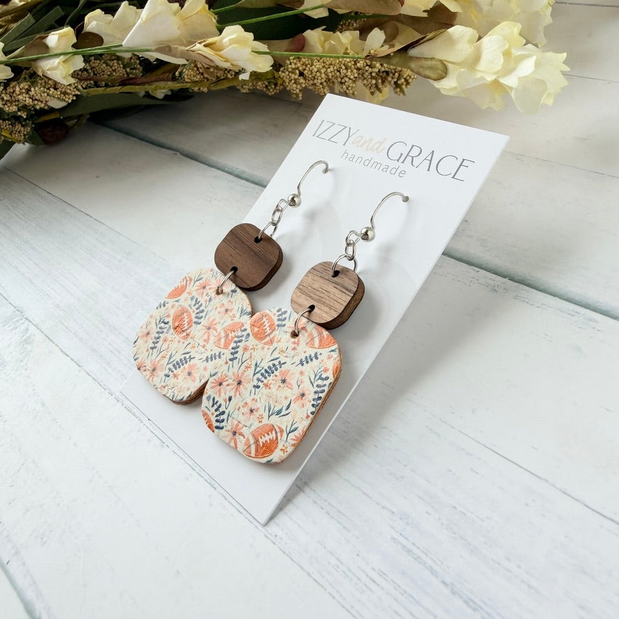 A pair of handmade dangle earrings displayed on a white card labeled 'Lizzy's Grace handmade.' The earrings have silver fishhook wires, small rectangular brown wooden tops, and larger cream-colored leather bottoms printed with a floral pattern in pink and blue.
