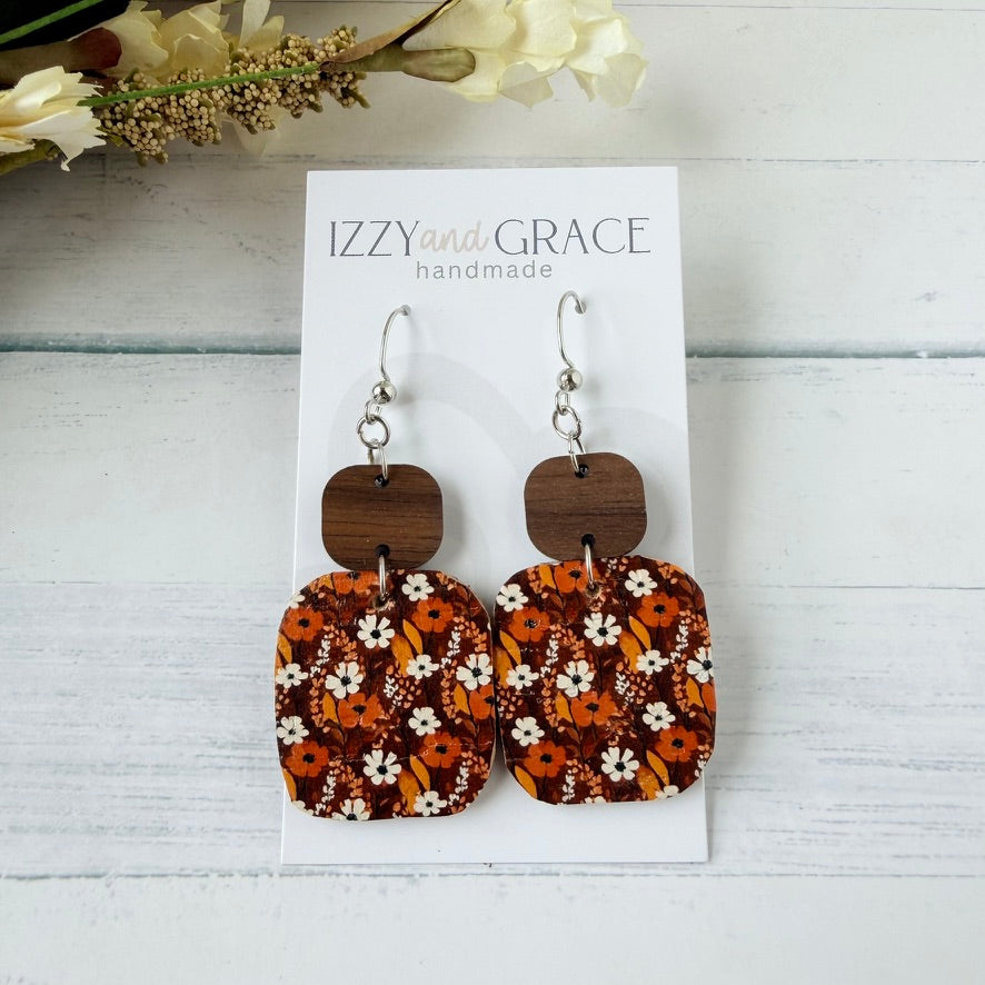 A pair of dangling earrings displayed on a white card labeled 'IZZYand GRACE handmade.' The earrings have silver hooks, dark brown wooden square tops, and rectangular lower pieces with an orange, brown, white, and black floral pattern.