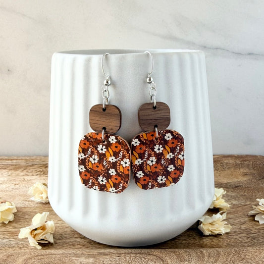 A pair of dangling earrings with rectangular floral panels in orange, brown, and white, attached to dark wood squares and silver hooks, displayed on a white ribbed ceramic mug.