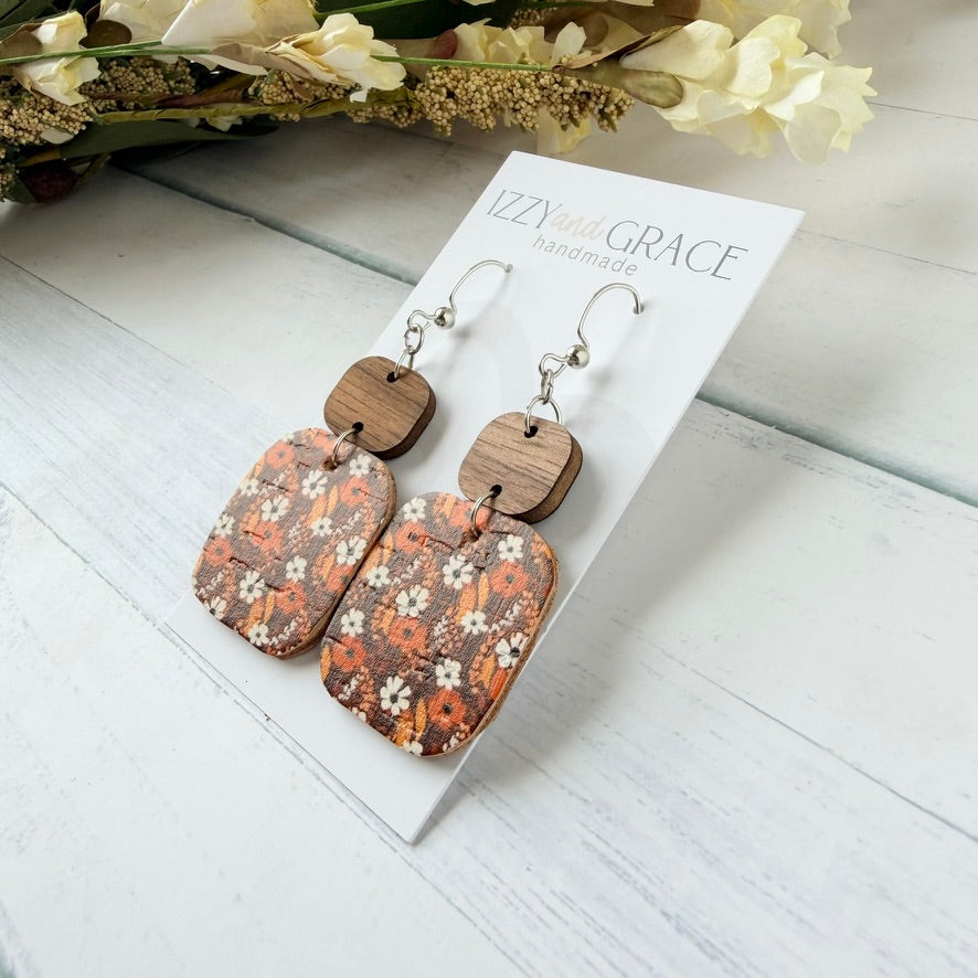 A pair of handmade drop earrings with rectangular floral-patterned wooden charms and silver hooks, displayed on a white card labeled 'Izzy Grace Handmade' on a white wood surface with dried flowers in the background.